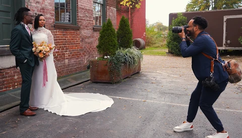 Wedding photographer capturing bride and groom posing beside a brick building during an outdoor portrait session.