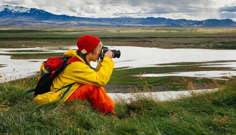 Photographer in yellow jacket and red hat crouching while photographing a river valley landscape with mountains.