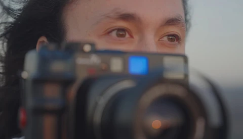 Photographer holding a professional video camera with blue display screen visible, shot through the camera lens with shallow depth of field.