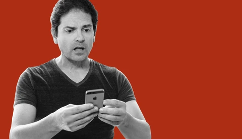 Man in black v-neck t-shirt holding a smartphone, speaking expressively against a red background.