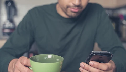 Man holding green cup and smartphone while looking at phone screen.