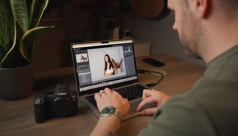 Photo editor working on a laptop displaying image editing software with a Canon camera and plant on wooden desk.