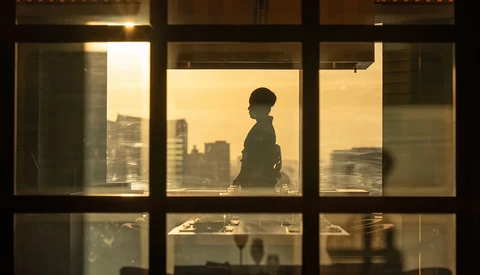 Silhouetted figure standing at a high-rise window overlooking a cityscape at golden hour.