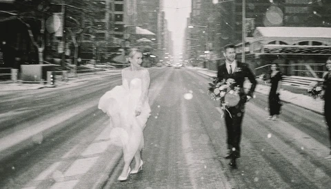 Bride in white dress running down urban street with groom and wedding party following behind.