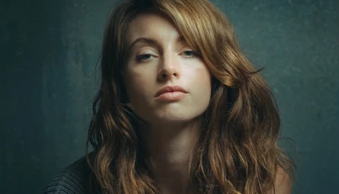 Portrait of a woman with long wavy brown hair against a muted teal background.