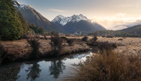 Snow-capped mountain peaks rise above a meandering stream in an expansive valley at golden hour.