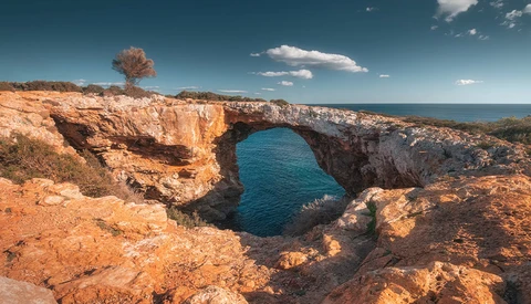 Natural rock arch formation with turquoise water visible through the opening, surrounded by russet cliffs under clear blue sky.