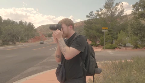 Photographer holding DSLR camera to eye while standing on roadside with mountains in background.