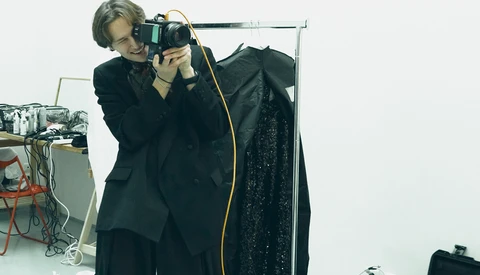 Photographer holding a camera to their eye while standing next to a clothing rack with a dark garment.