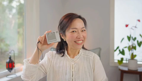 Woman holding a compact digital camera toward the camera with a bright smile indoors.