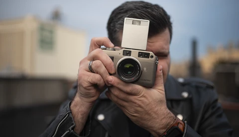 Photographer holding a rangefinder camera with flash attachment to his face while composing a shot.