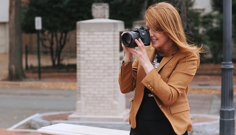 Woman in tan blazer holding a digital camera to her face while standing in an urban plaza.