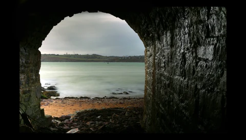 Seascape framed through a stone tunnel opening, showing turquoise water and distant shoreline.