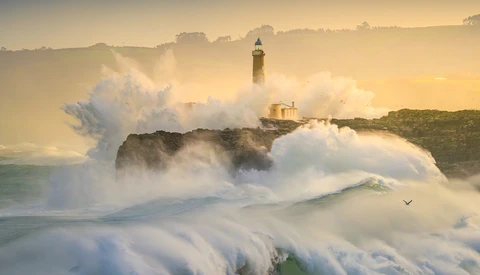 Lighthouse on rocky headland surrounded by massive crashing waves and sea spray at sunrise.