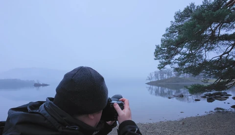 Photographer shooting landscape at dawn by a misty lake with pine trees reflected in still water.