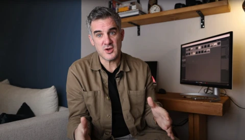 Man in tan jacket speaking to camera in home studio with monitor displaying editing software.
