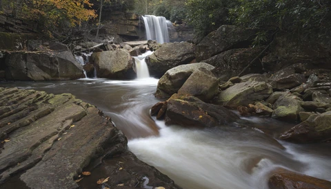 Cascading waterfall flowing over moss-covered rocks into a misty stream surrounded by dense forest.