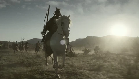 Silhouetted rider on horseback leading a group across an arid, mountainous desert landscape at sunset.