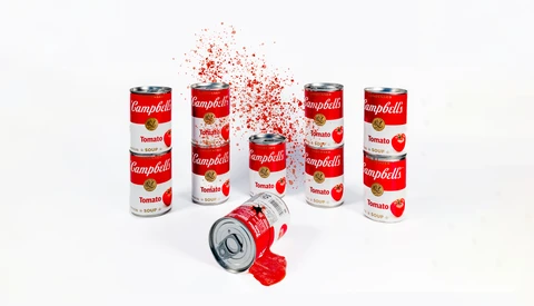Campbell's tomato soup cans arranged in a group with one tipping over and dispersing red particles into the air.