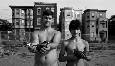 Two shirtless boys holding small animals in an urban lot with rowhouses behind them.