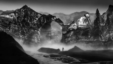 Solitary figure standing in misty valley surrounded by dramatic rocky peaks in black and white.