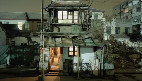 Weathered two-story residential building with illuminated doorway at night, surrounded by older structures.