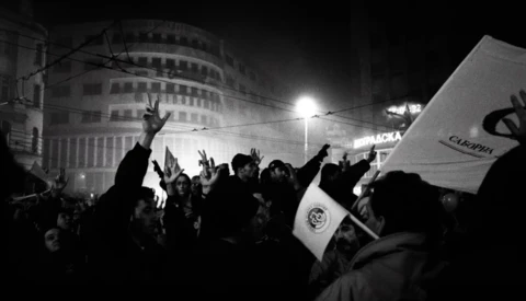 Nighttime crowd protest with raised hands and flags, urban buildings lit by street lamps.