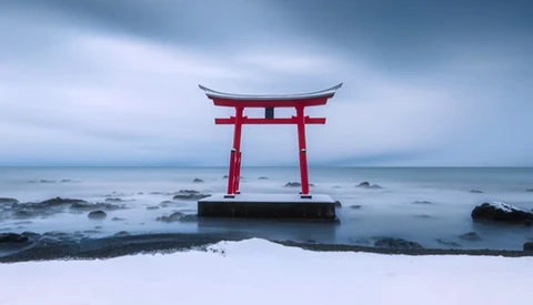Red torii gate standing in shallow ocean water with snow-covered beach and rocky coastline.