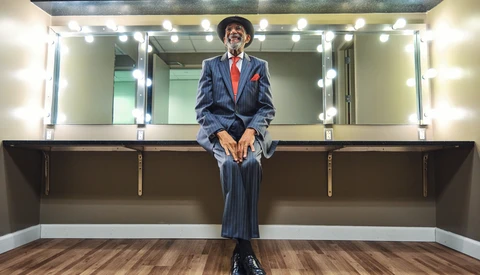Person in blue pinstriped suit and red tie sitting on vanity counter beneath illuminated mirror with multiple bulbs.