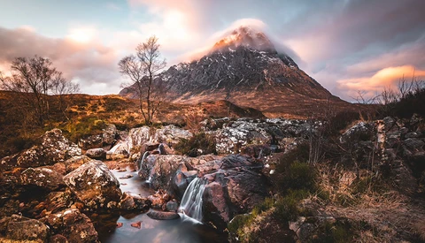 Snow-capped mountain peak at sunset with a small waterfall flowing over rocks in the foreground.