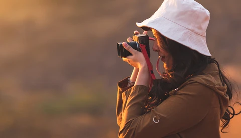 Photographer with white bucket hat holding camera to face, backlit by warm golden light against blurred background.