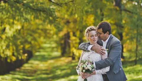 Groom embracing bride from behind in tree-lined garden setting during golden hour.