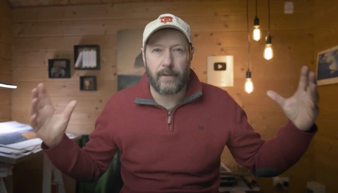Man in burgundy quarter-zip pullover and white cap gestures expressively while speaking in a warm, wood-paneled interior space.