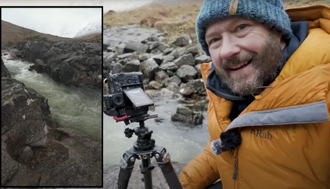 Filmmaker smiling at camera while operating cinema camera on tripod beside rushing glacial river in rocky mountain valley.