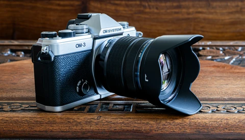 OM System OM-3 film camera with black leather grip and attached wide-angle lens with hood on wooden surface.
