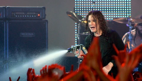 Performer on stage with long dark hair singing into microphone, wearing black jacket over red shirt, with amplifier and LED screen backdrop.