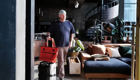 Man standing in a loft studio space holding a red bag next to packed luggage and moving boxes.