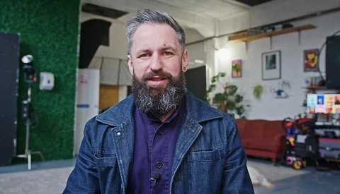 Bearded man in denim jacket smiling at camera in creative studio space.
