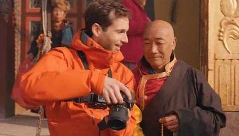 Man in orange jacket holding binoculars speaks with elderly man wearing brown robe and prayer beads.