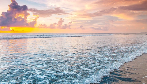 Tropical beach at sunset with golden-hour sky, gentle waves, and dramatic cloud formations.