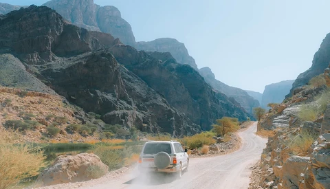 White SUV driving on a dusty mountain road through a dramatic canyon with steep rocky cliffs.