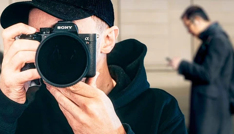 Photographer holding Sony mirrorless camera up to face while wearing black cap and jacket.