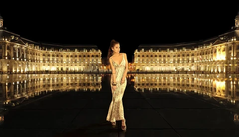 Woman in an elegant beaded gown standing centered in a symmetrical plaza reflected in still water at night.