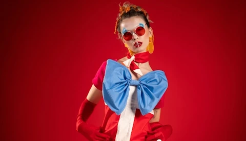 Woman in red jacket and blue bow tie against vibrant red background wearing round sunglasses.