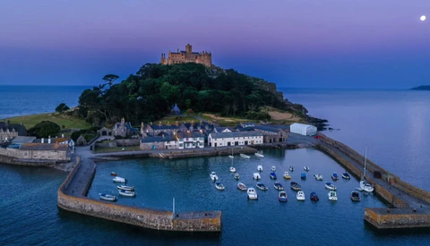 Coastal village with castle on hilltop at twilight, moored boats in harbor below.