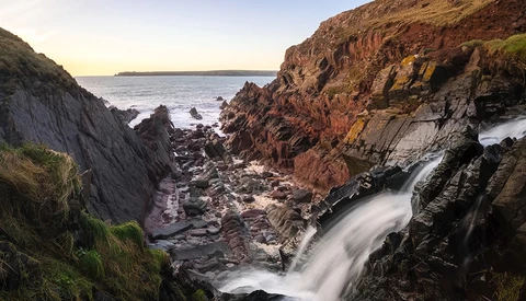 Coastal rock formation with waterfall and long-exposure ocean waves at sunrise.