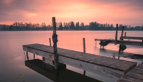 Wooden boat docks extending into a calm lake at sunrise, with pink sky and forested shoreline.