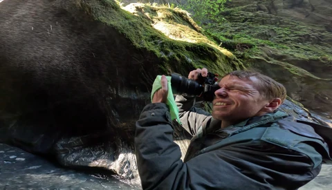 Photographer holding a DSLR camera while crouching in a mossy canyon, smiling while composing a shot.