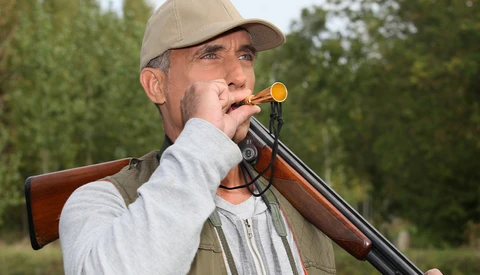 Hunter in cap and vest holding rifle with scope to shoulder, aiming outdoors.