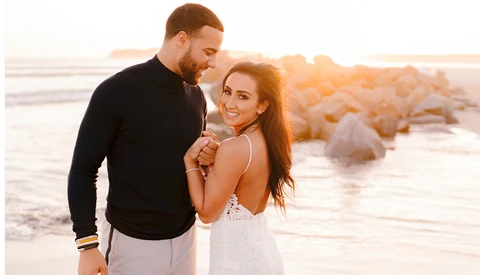 Couple smiling together on a beach at golden hour with rocky formations in the background.